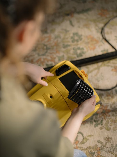 A person holds a yellow portable fan or air mover over a patterned carpet in a room with a textured brick wall. The device has a black grille and a digital display, suggesting use in surface cleaning or drying processes. The room is illuminated with natural or artificial light, reflecting a clean and organized environment. Visible is a black cable extending from the device, indicating it is plugged in for operation. The setting exemplifies professional cleaning equipment used by Bayswater Cleaners for enhancing hygiene and cleanliness, aligned with expertise in deep cleaning and surface sanitisation on the page titled 'Affordable carpet cleaning Porchester Road Bayswater prices'.