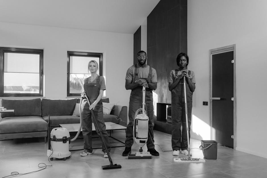 Three professional cleaners from Bayswater Cleaners are conducting surface cleaning and deep cleaning in a modern living room. The room features a large L-shaped sofa with a fabric upholstery, positio