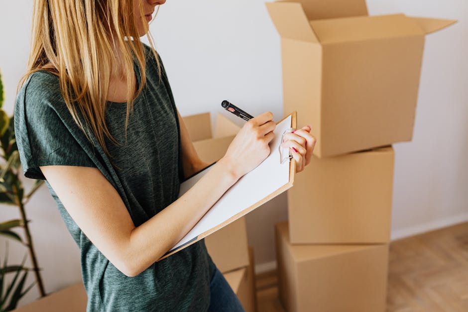 A woman with blonde hair, wearing a dark green T-shirt, is seen holding a clipboard and a black marker, preparing to take notes or check off items. Behind her, there are several stacked cardboard boxes, some open, on a wooden floor in a well-lit room with white walls. The scene suggests an end-of-tenancy or moving process, related to professional cleaning or preparation for property inspection. The background includes a green leafy plant, adding a touch of decor to the space, and the overall setting emphasizes cleanliness and organization. Bayswater Cleaners provides detailed surface cleaning, deep cleaning, and sanitisation services suitable for moving out or end-of-tenancy cleaning, as highlighted on their webpage 'End of tenancy cleaning Bayswater W2 quote and checklist'.