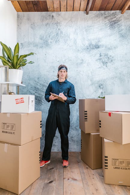 A woman dressed in dark blue work overalls and a headscarf holding a clipboard stands in a room with a industrial-style concrete wall and wooden ceiling beams. She is positioned among several packed cardboard boxes with printed labels, indicating recent or ongoing move or storage activities. To her left, there is a white shelf holding a potted plant, which adds a touch of greenery to the scene. The room has natural lighting that highlights the clean and organized appearance of the space, and the wooden floor beneath her complements the warm, inviting atmosphere. This setting reflects professional domestic and commercial cleaning services offered by Bayswater Cleaners, emphasizing thorough surface cleaning, organisation, and preparation for end-of-tenancy or deep cleaning tasks.