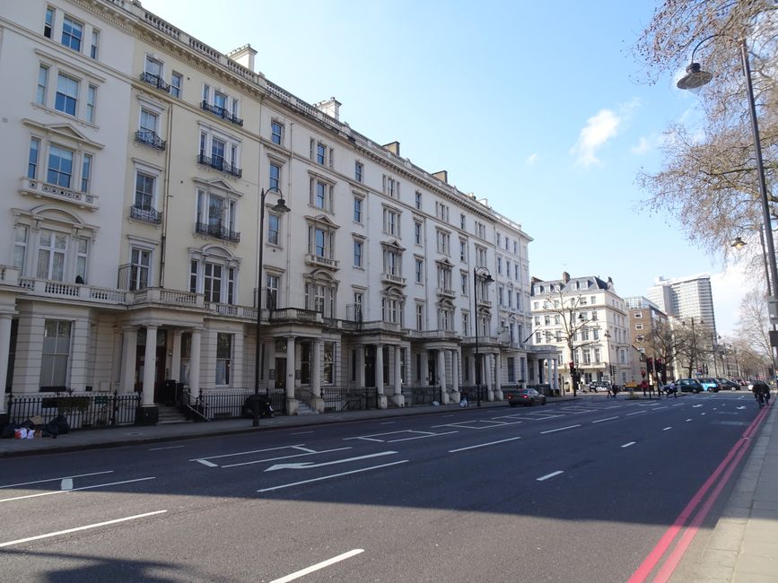 A view of a classic white Victorian-style apartment building along a city street, showcasing detailed architectural features such as decorative window frames, stone columns at the entrance, and wrought iron balconies. The building's facade appears clean and well-maintained under bright daylight, with clear blue skies overhead. The street in front has marked lanes and parked cars, with leafless trees lining the sidewalk and a street lamp visible on the right. The scene reflects urban cleanliness and upkeep, representative of professional domestic or commercial cleaning standards that Bayswater Cleaners offers for residential and commercial properties in the Hyde Park area.