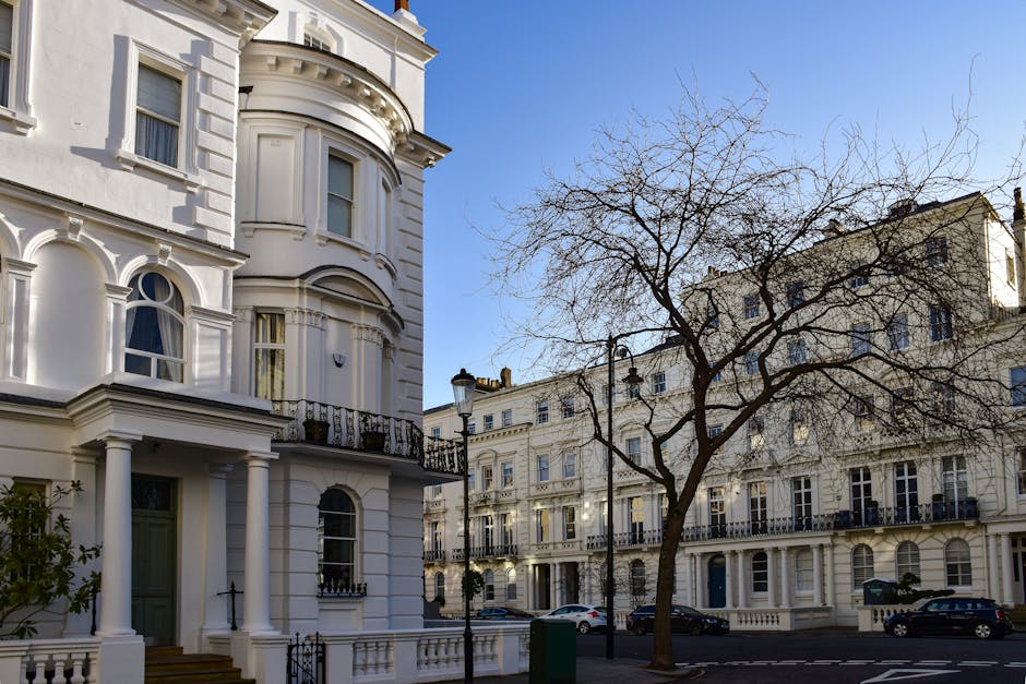 View of a residential street in Bayswater with classic white Victorian-style terraced houses featuring decorative mouldings and large sash windows, and a prominent corner building with rounded bay windows and columns at the entrance. A leafless tree stands in the foreground alongside street lamps and parked cars under a clear blue sky, illustrating a clean and well-maintained urban environment. This exterior scene reflects the aesthetic typical of Hyde Park view flats and highlights the importance of ongoing surface cleaning and maintenance, as reinforced by Bayswater Cleaners' domestic cleaning and sanitisation services.