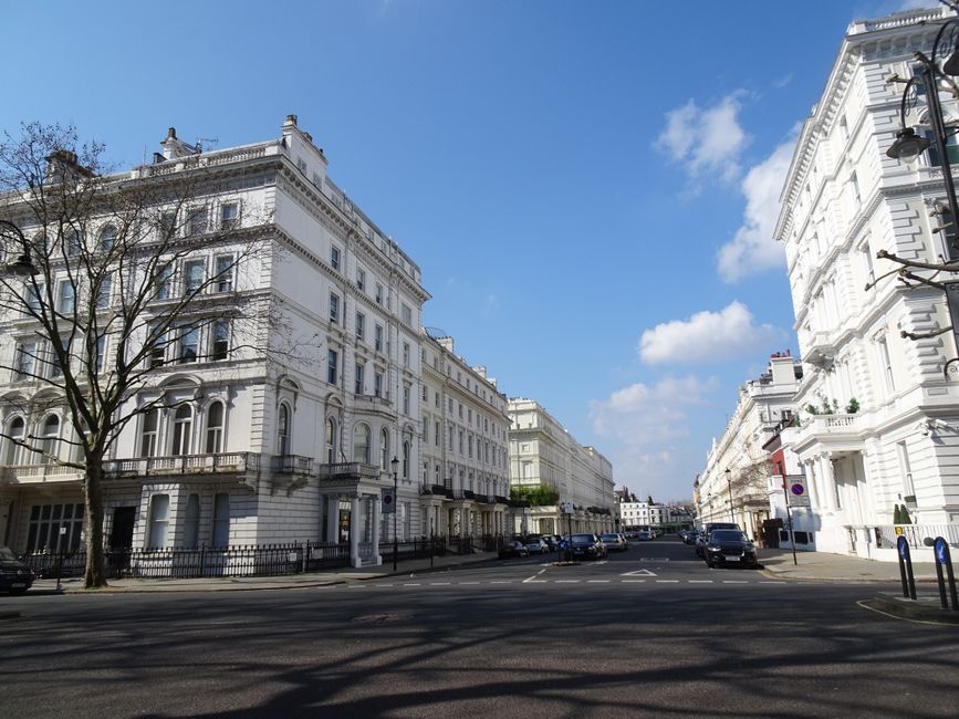 A view of a classic white Victorian-style apartment building along a city street, showcasing detailed architectural features such as decorative window frames, stone columns at the entrance, and wrought iron balconies. The building's facade appears clean and well-maintained under bright daylight, with clear blue skies overhead. The street in front has marked lanes and parked cars, with leafless trees lining the sidewalk and a street lamp visible on the right. The scene reflects urban cleanliness and upkeep, representative of professional domestic or commercial cleaning standards that Bayswater Cleaners offers for residential and commercial properties in the Hyde Park area.
