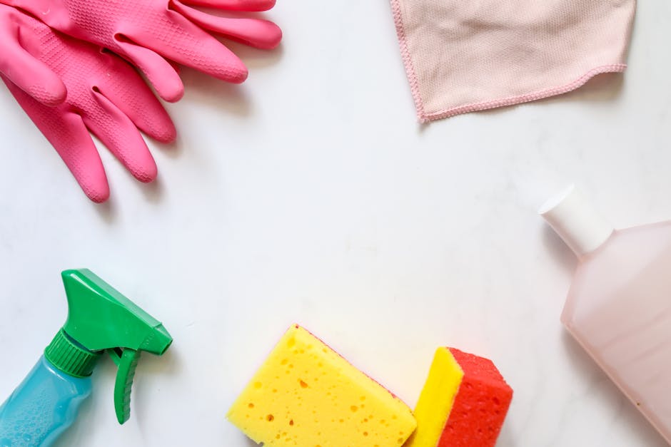An overhead view of cleaning supplies arranged on a white surface including a pair of pink rubber gloves, a pink cloth, a green spray bottle, a yellow and red sponge, and a white plastic bottle with a cap. The items are placed in a way that suggests preparation for surface cleaning or sanitisation in a domestic or commercial setting. The natural lighting highlights the clean and organized appearance of the cleaning tools, emphasizing the importance of thorough surface cleaning and hygiene. Bayswater Cleaners recommends proper use of these supplies for effective deep cleaning and maintaining a hygienic environment, as outlined in their Queensway flat cleaning tips for landlords and tenants.