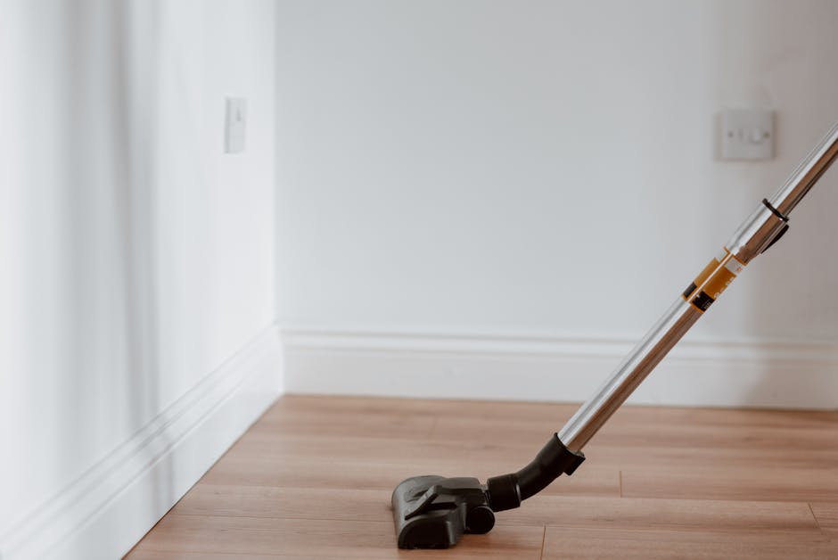A vacuum cleaner with a black floor head and a long, metallic silver and black wand is positioned on a light wooden floor in a well-lit room. The background features a white wall with two electrical outlets near the baseboard. The scene depicts surface cleaning and maintenance, emphasizing the effectiveness of equipment used by Bayswater Cleaners in deep cleaning and sanitisation processes for residential spaces, specifically in a flat setting as part of their domestic cleaning services.